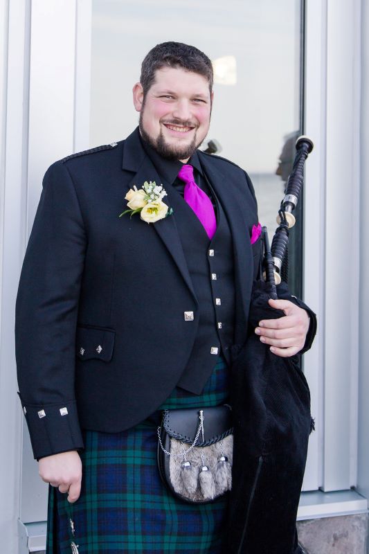 Aaron dressed up in formal attire - black vest and jacket with a kilt and sporran. He's holding bagpipes